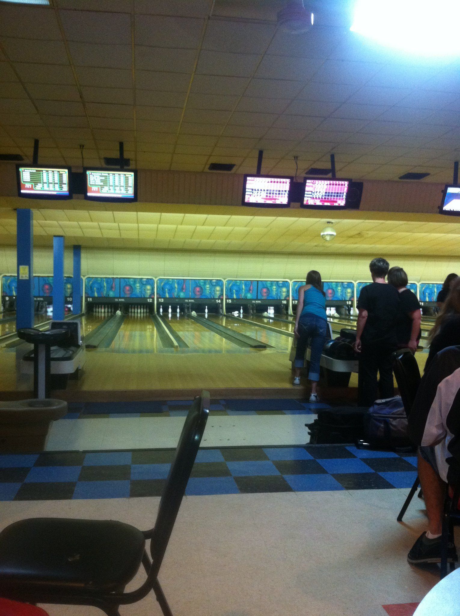 People preparing to bowl at Oil Bowl Lanes with active score monitors and blue checkered flooring in the foreground.