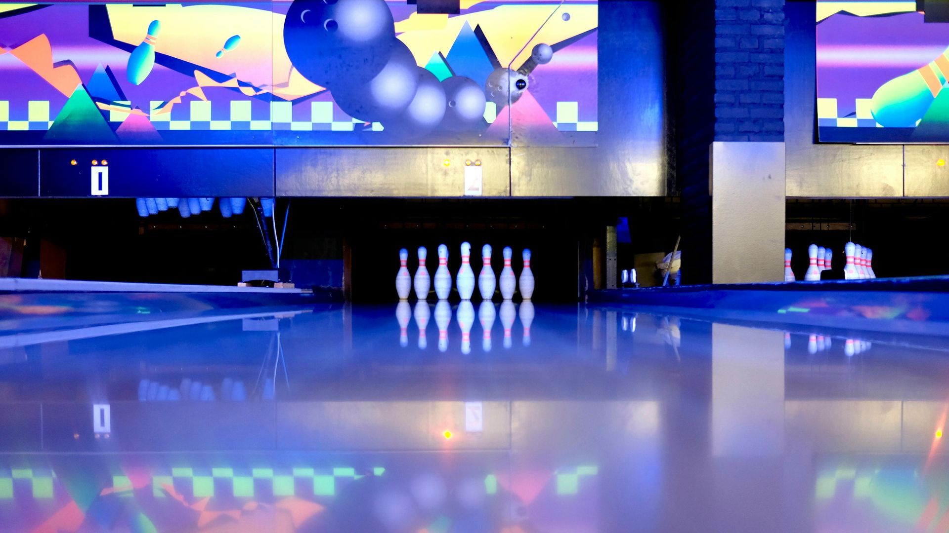 A low-angle view of white bowling pins set on a reflective blue lane under neon cosmic lighting.