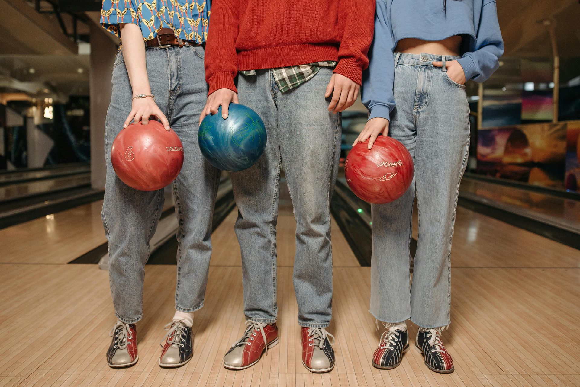 Three people in denim jeans and retro bowling shoes hold bowling balls while standing on a lane.