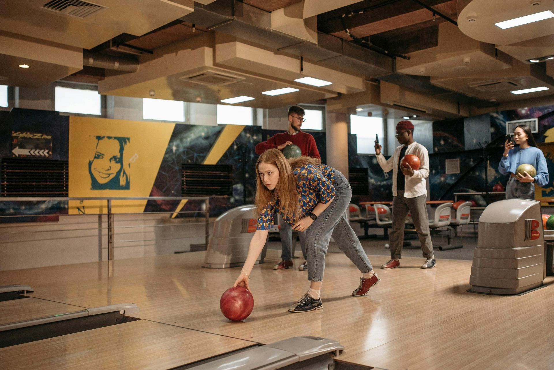 A young woman releases a red bowling ball on a lane while her friends watch from behind.