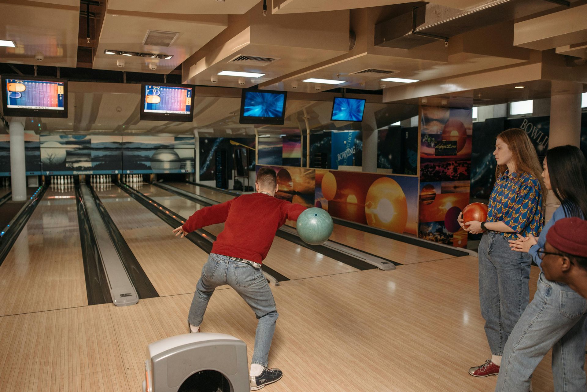 A bowler in a red sweater releases a ball down a lane while three friends watch from the side.