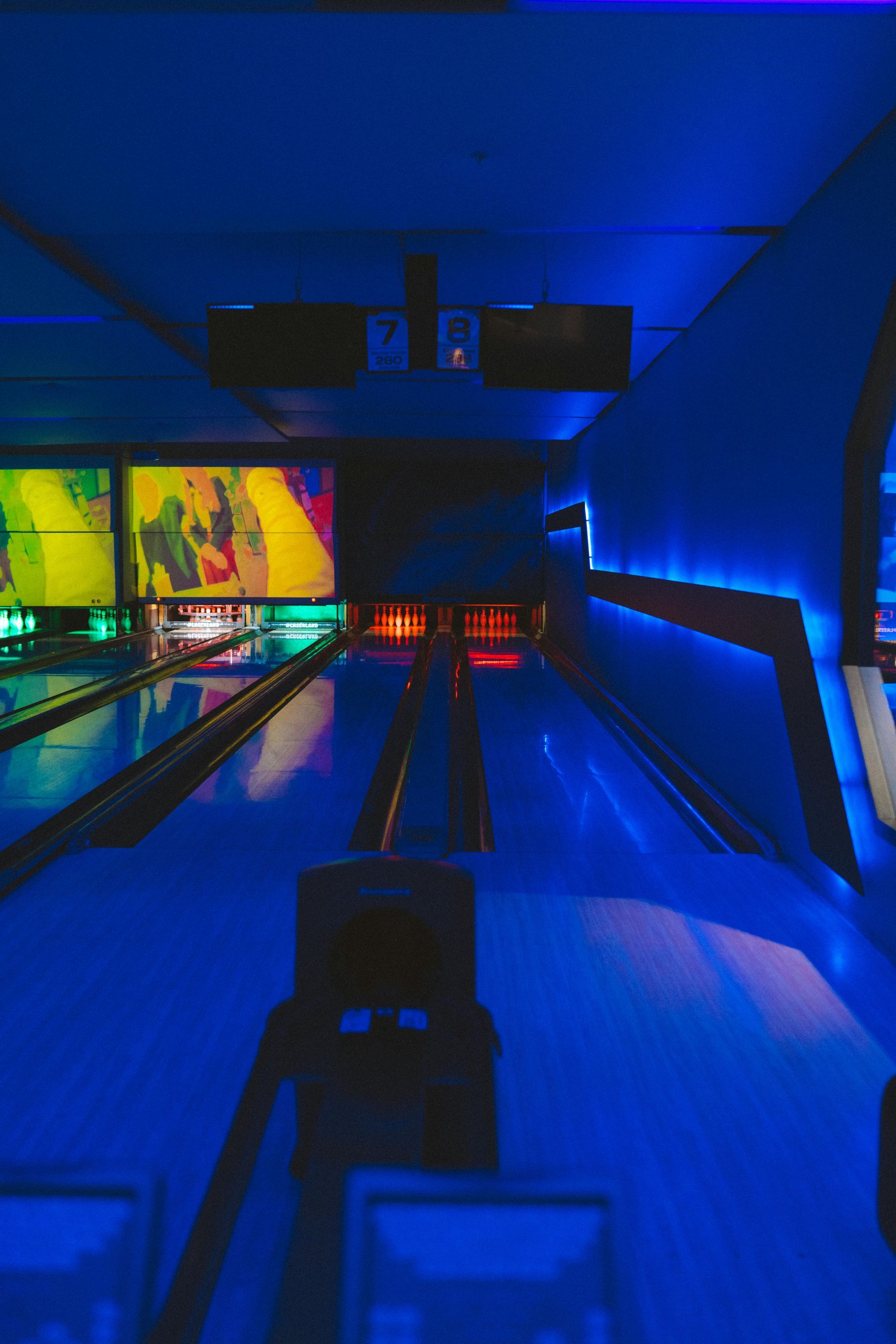 View down two bowling lanes under vibrant blue cosmic lighting with glowing pins and wall accents.