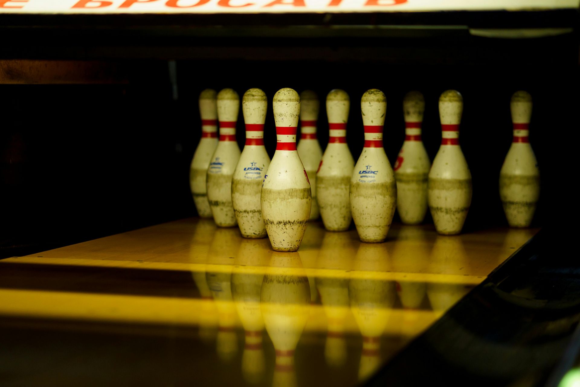 Ten bowling pins stand in formation at the end of a polished wooden lane with a clear reflection.