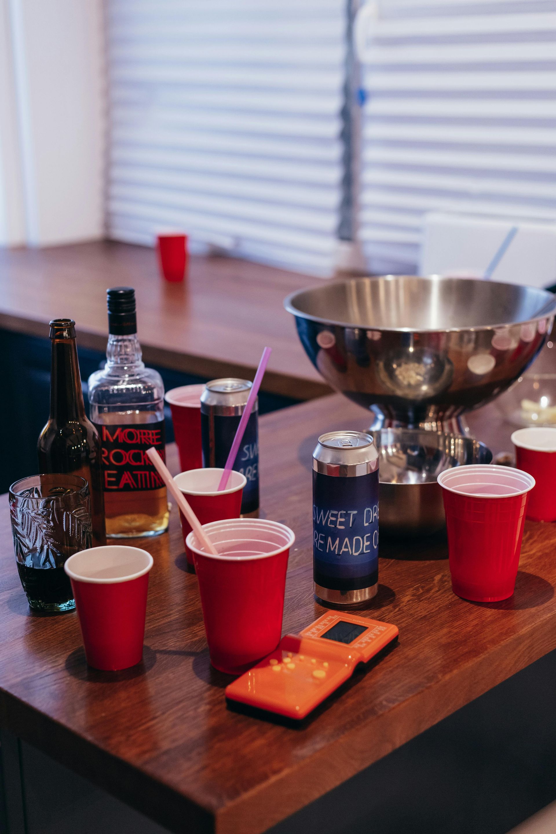 A wooden bar counter featuring red cups, a liquor bottle, drink cans, a metal bowl, and an orange handheld game console.