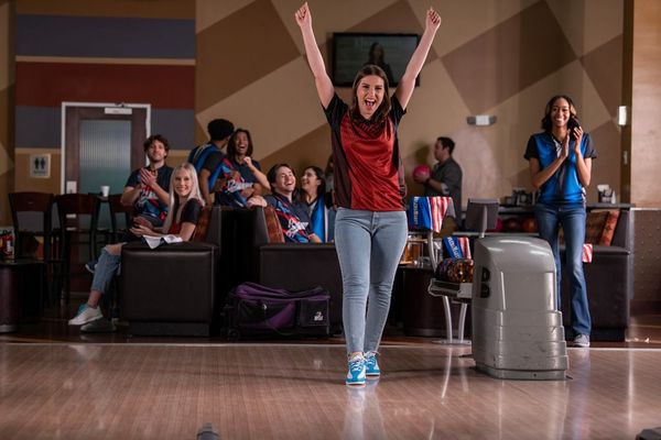 A woman in a bowling jersey celebrates with her arms raised while friends cheer behind her at a bowling alley.
