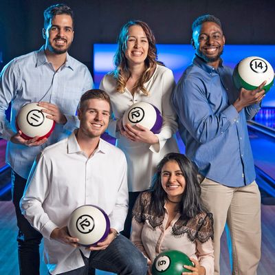 Five smiling people holding bowling balls designed to look like giant billiard balls at a bowling alley.