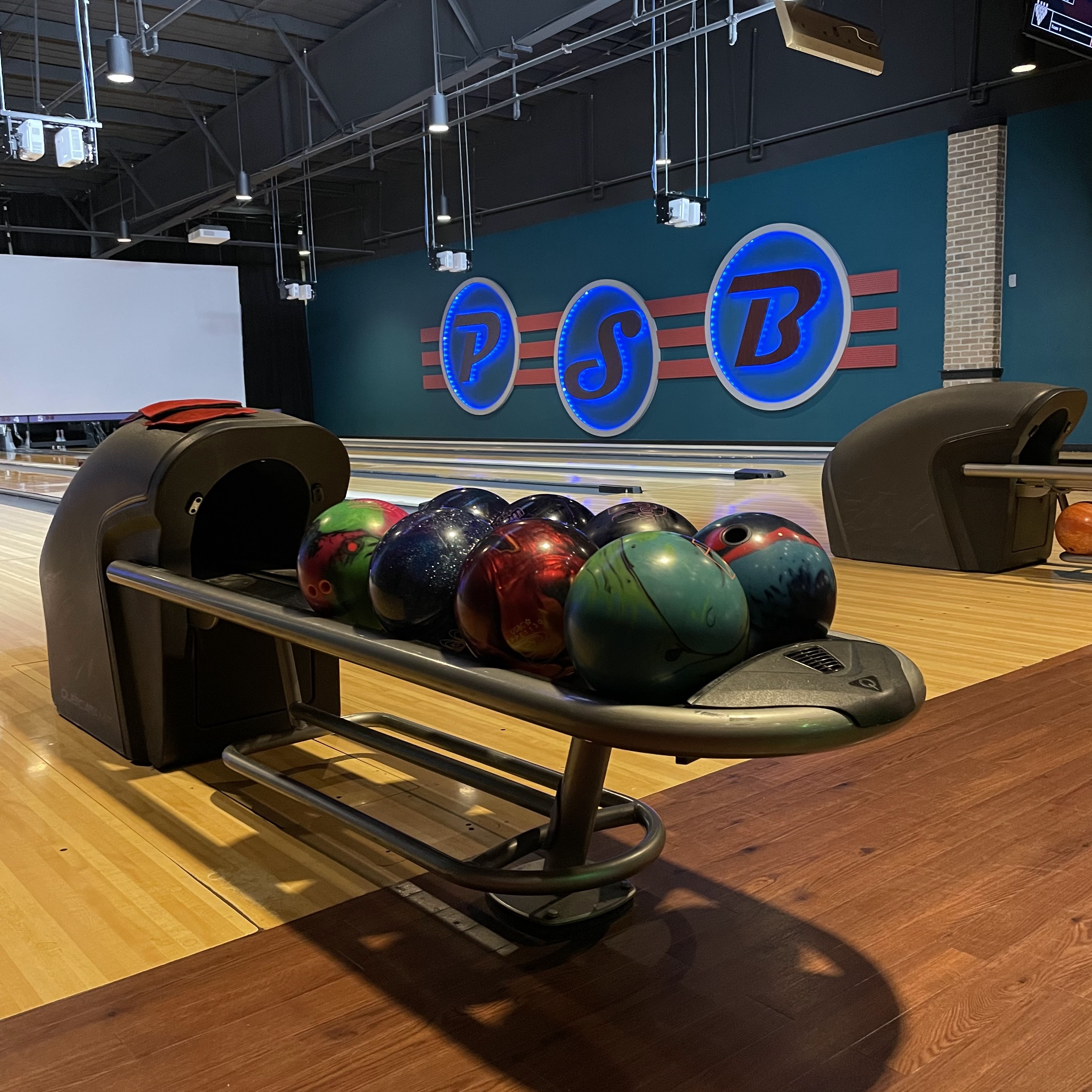 A variety of colorful bowling balls sit on a sleek ball return in front of a wall with a glowing PSB logo.