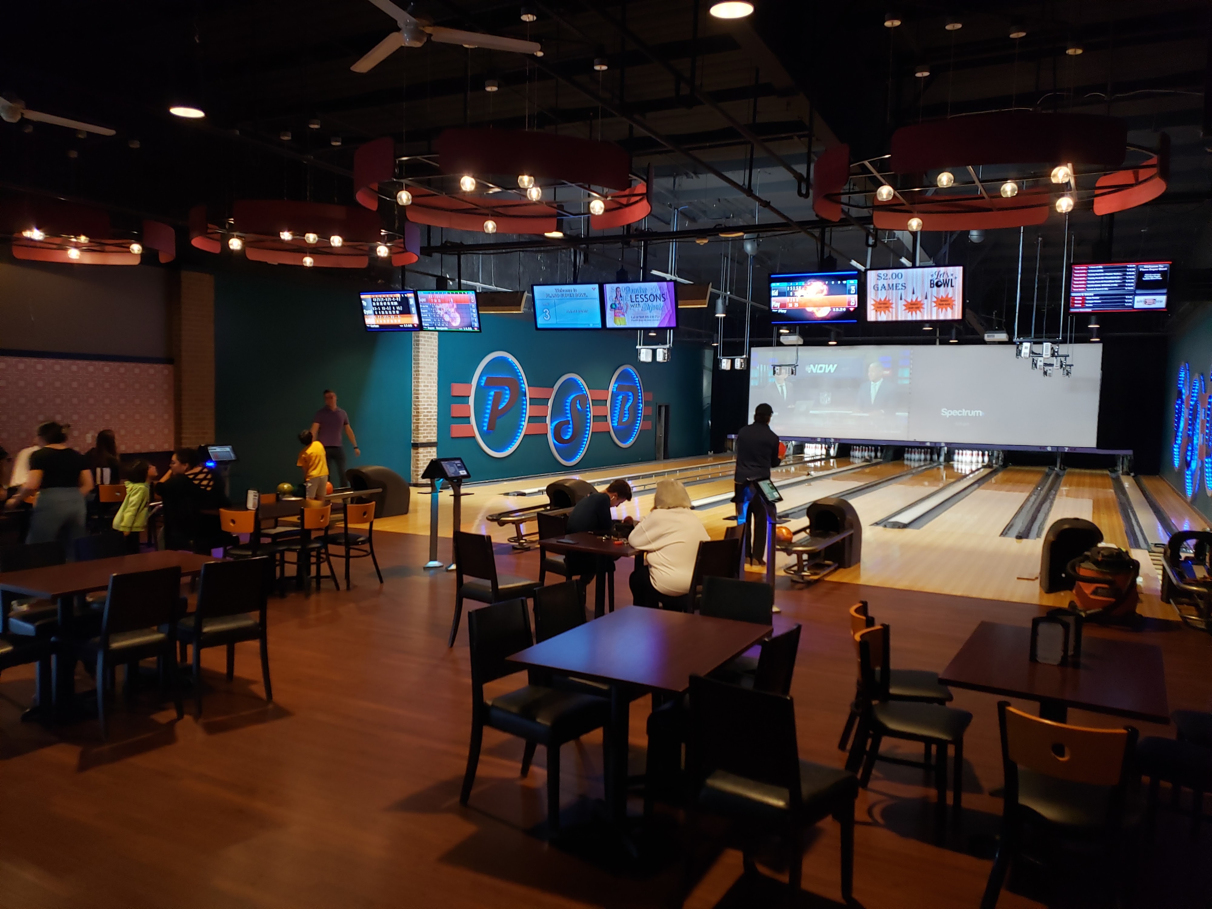 Interior view of a bowling center showing lanes, a large projection screen, and a dining area with tables and chairs.