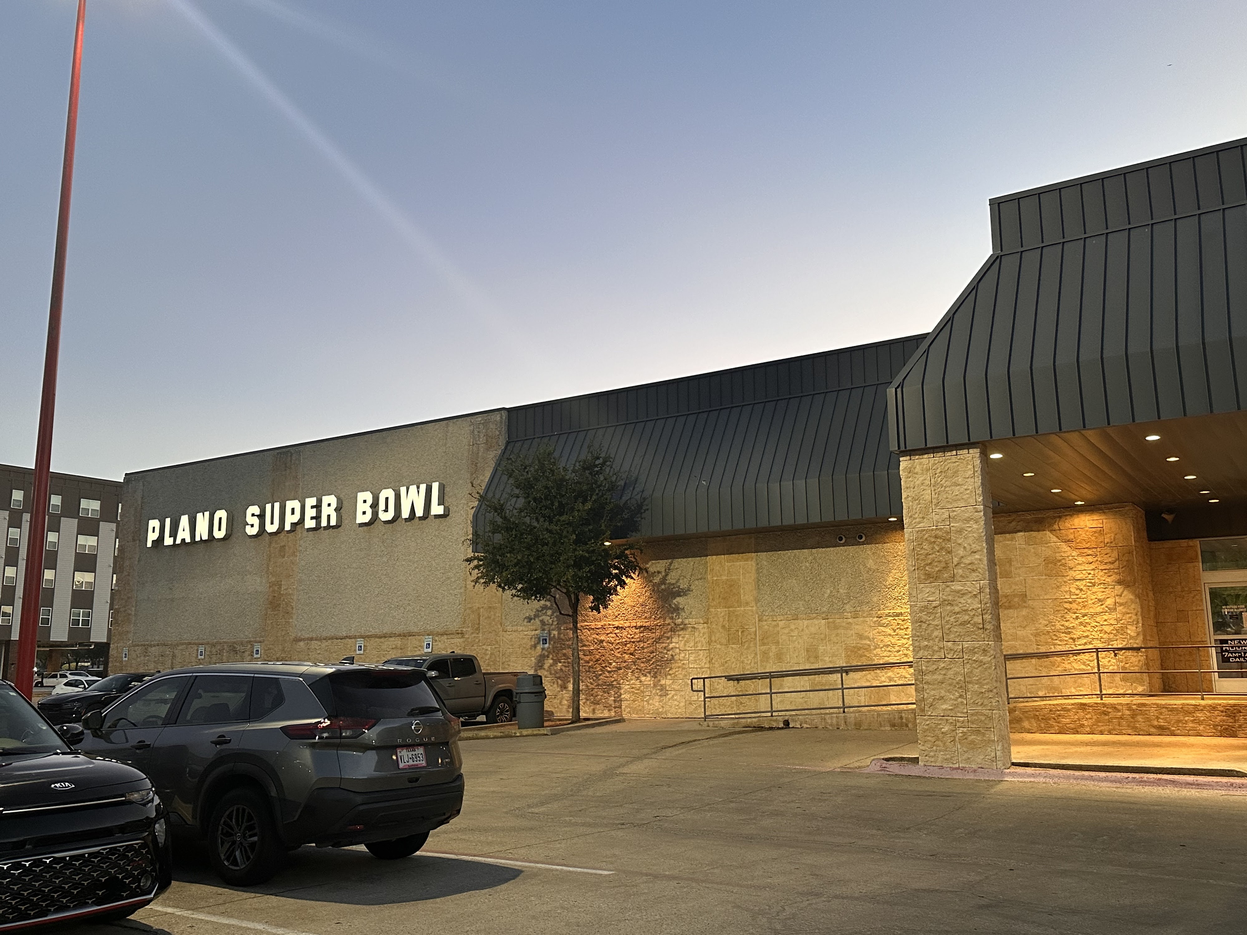 Exterior view of the Plano Super Bowl building at dusk with a large parking lot and lighted entrance.