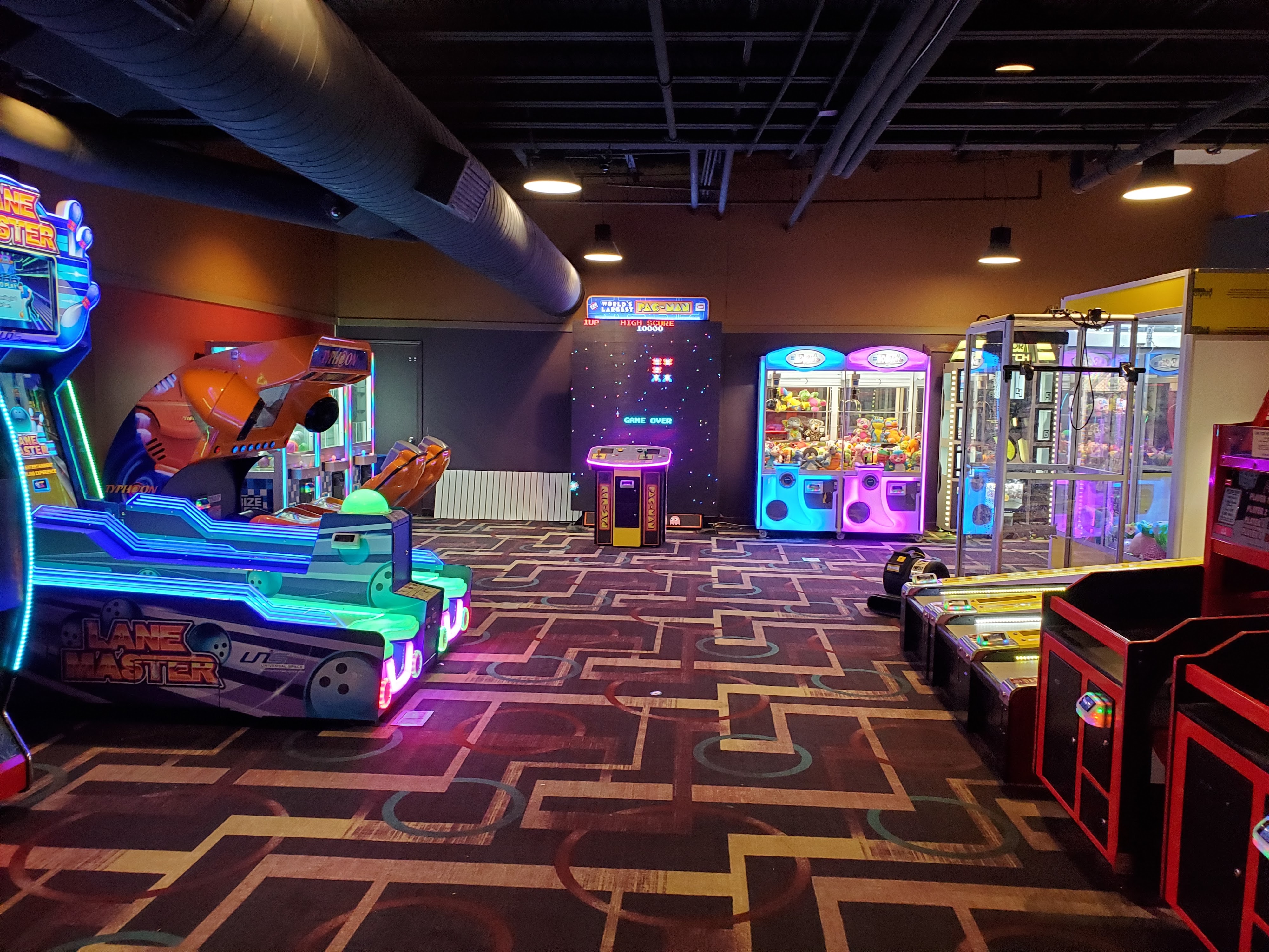 A brightly lit arcade room with various video games and prize crane machines on a patterned carpet.