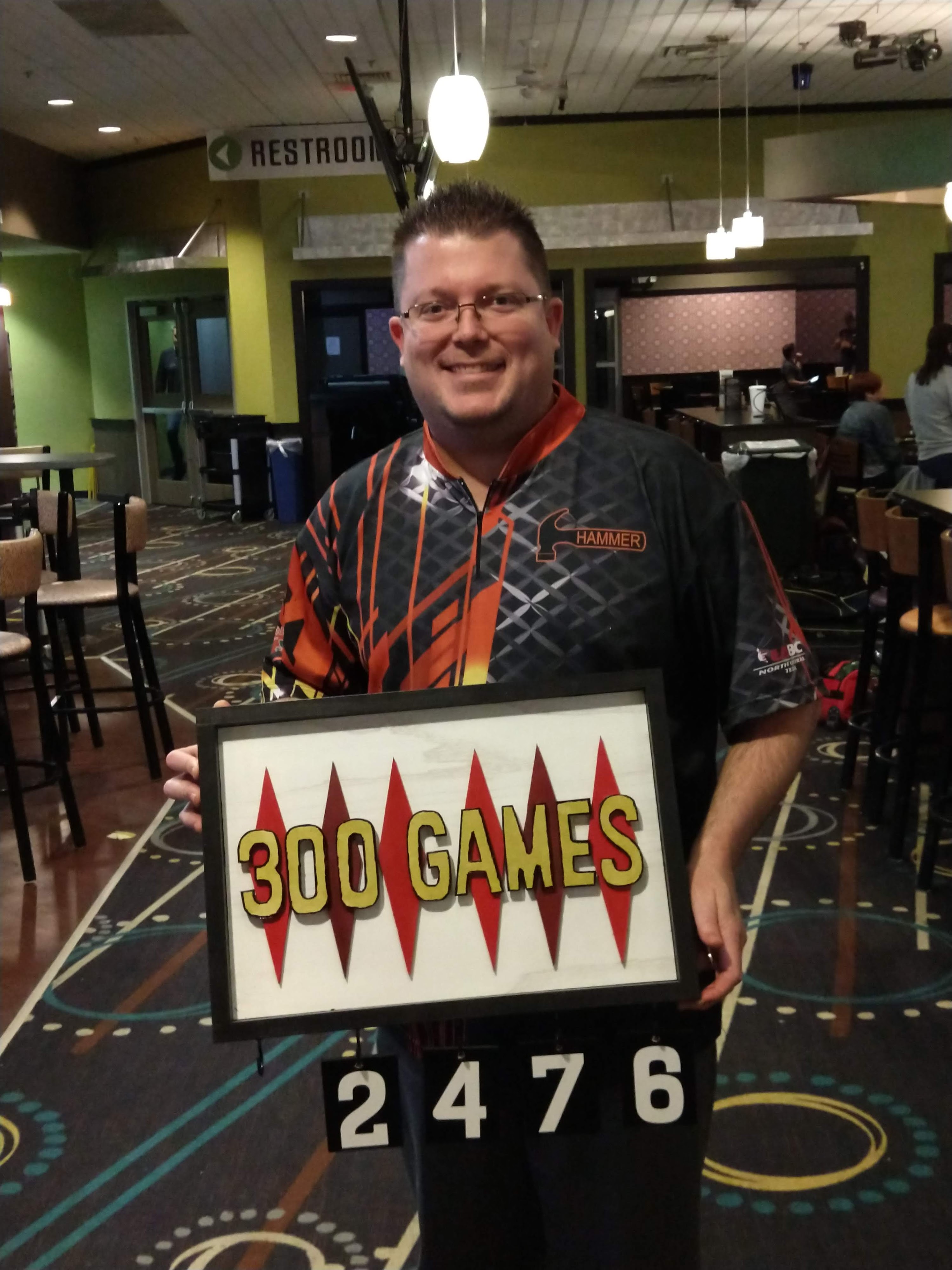 A man in a bowling jersey smiling while holding a framed sign that celebrates 300 games.