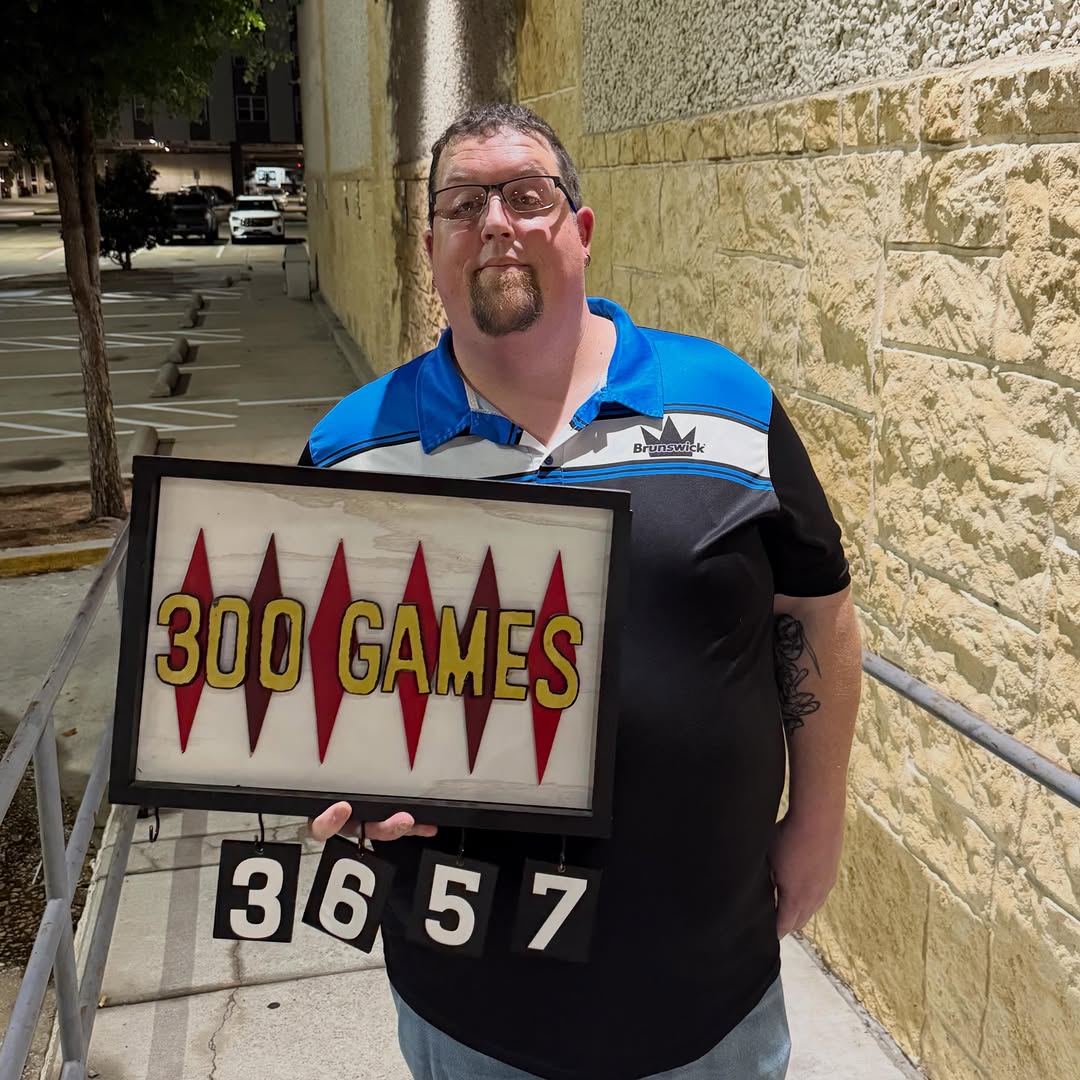 A man in a bowling jersey holds a 300 Games milestone sign outside at night.
