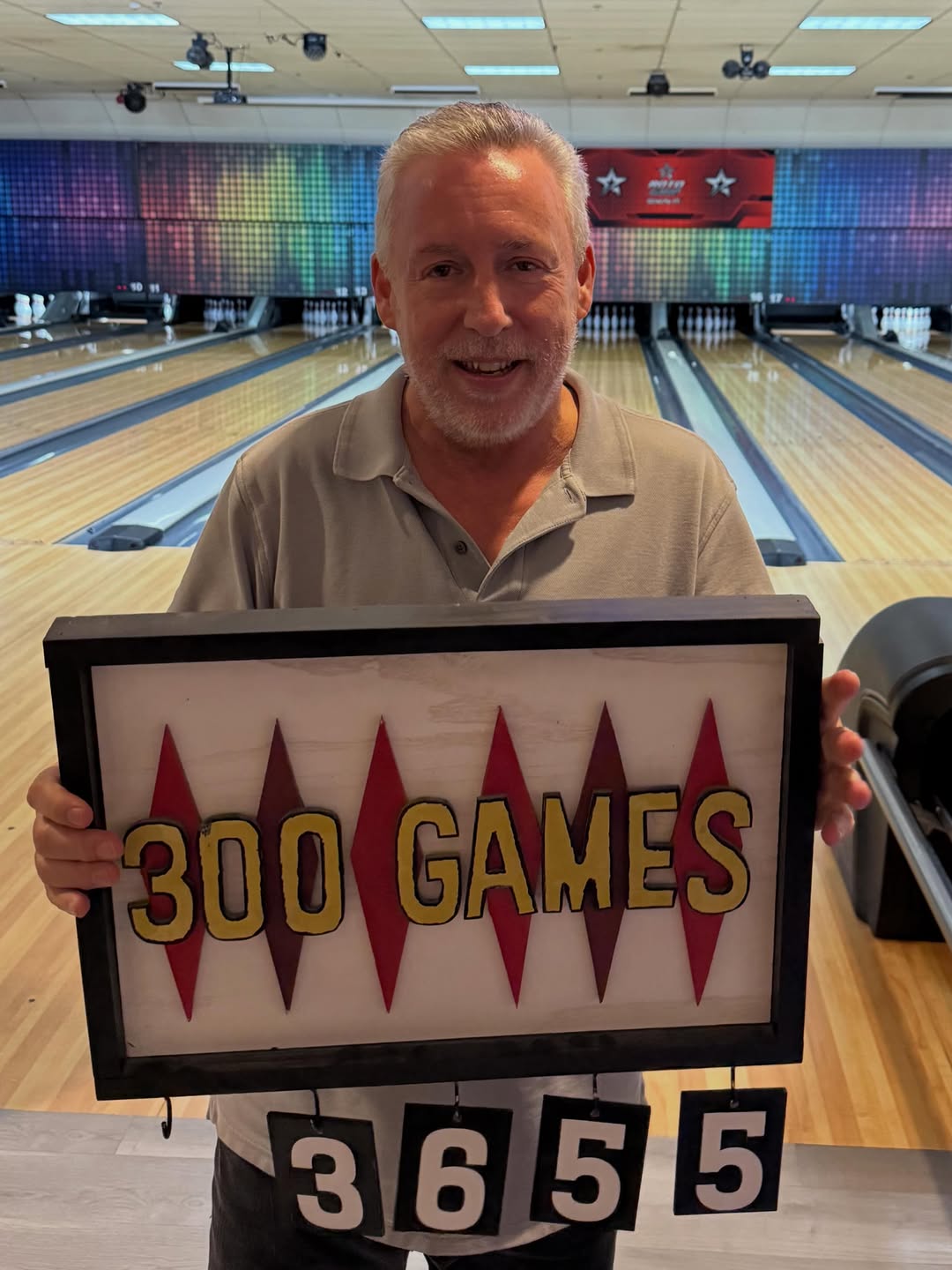 A man holding a 300 Games sign with the number 3655 on bowling lanes.
