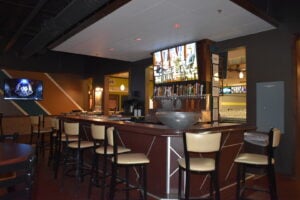 A modern bowling center bar area with high-back stools and televisions showing sports.