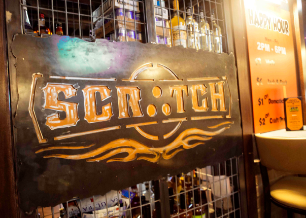 A rustic metal sign for Scratch Bar mounted over a liquor shelf next to a Happy Hour menu board.