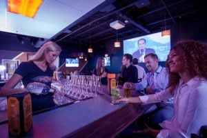 A bartender pours a drink for guests at a modern bar with large television screens in the background.