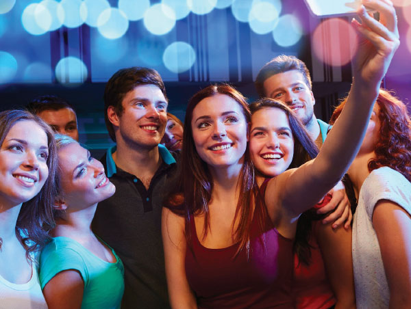 A group of young adults smiling for a selfie together in front of glowing blue and pink lights.