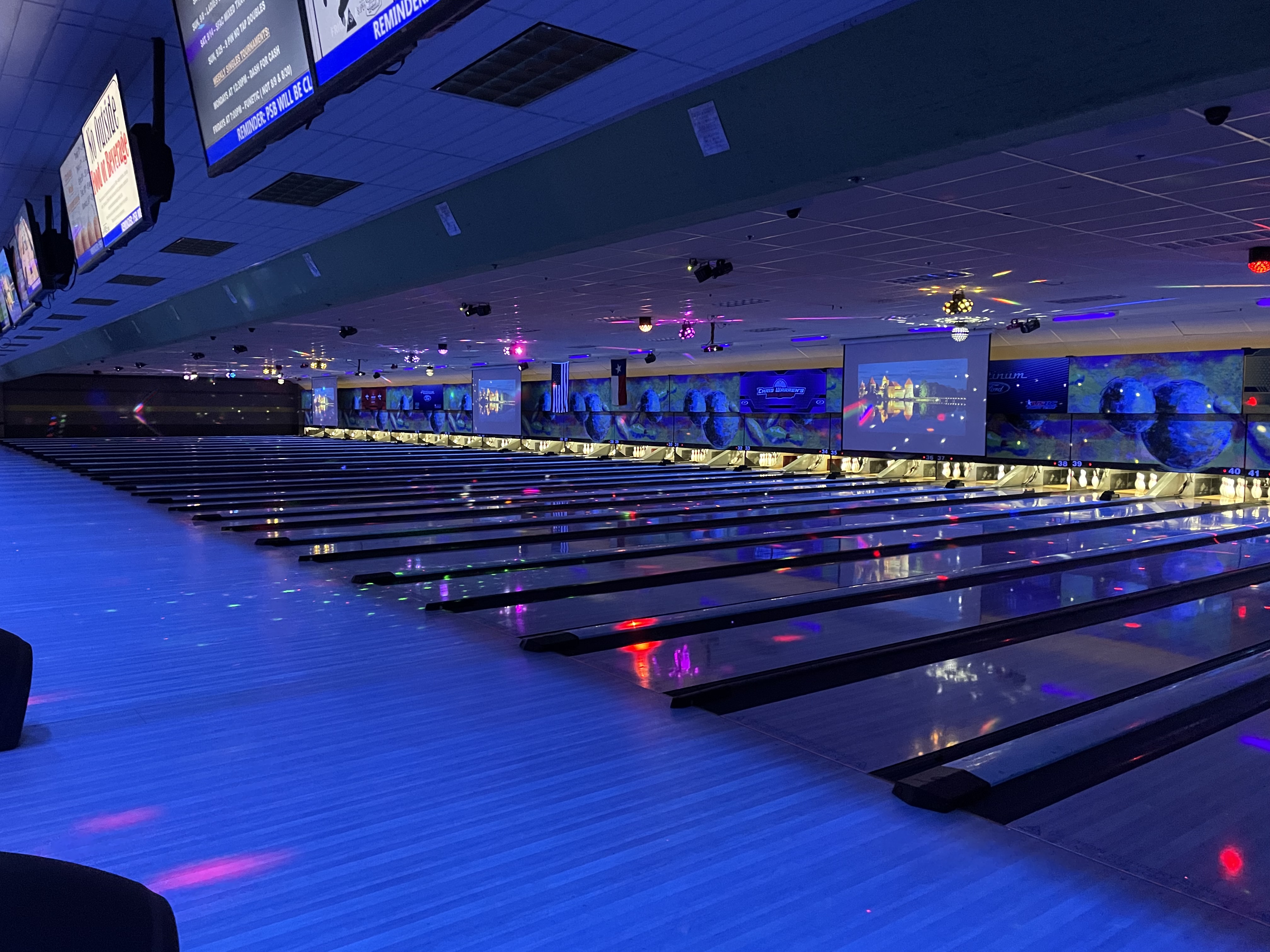 A wide row of bowling lanes illuminated with blue cosmic lighting and colorful neon reflections on the floor.