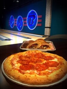 A close-up of a pepperoni pizza and a sliced calzone in front of bowling lanes and a neon logo.