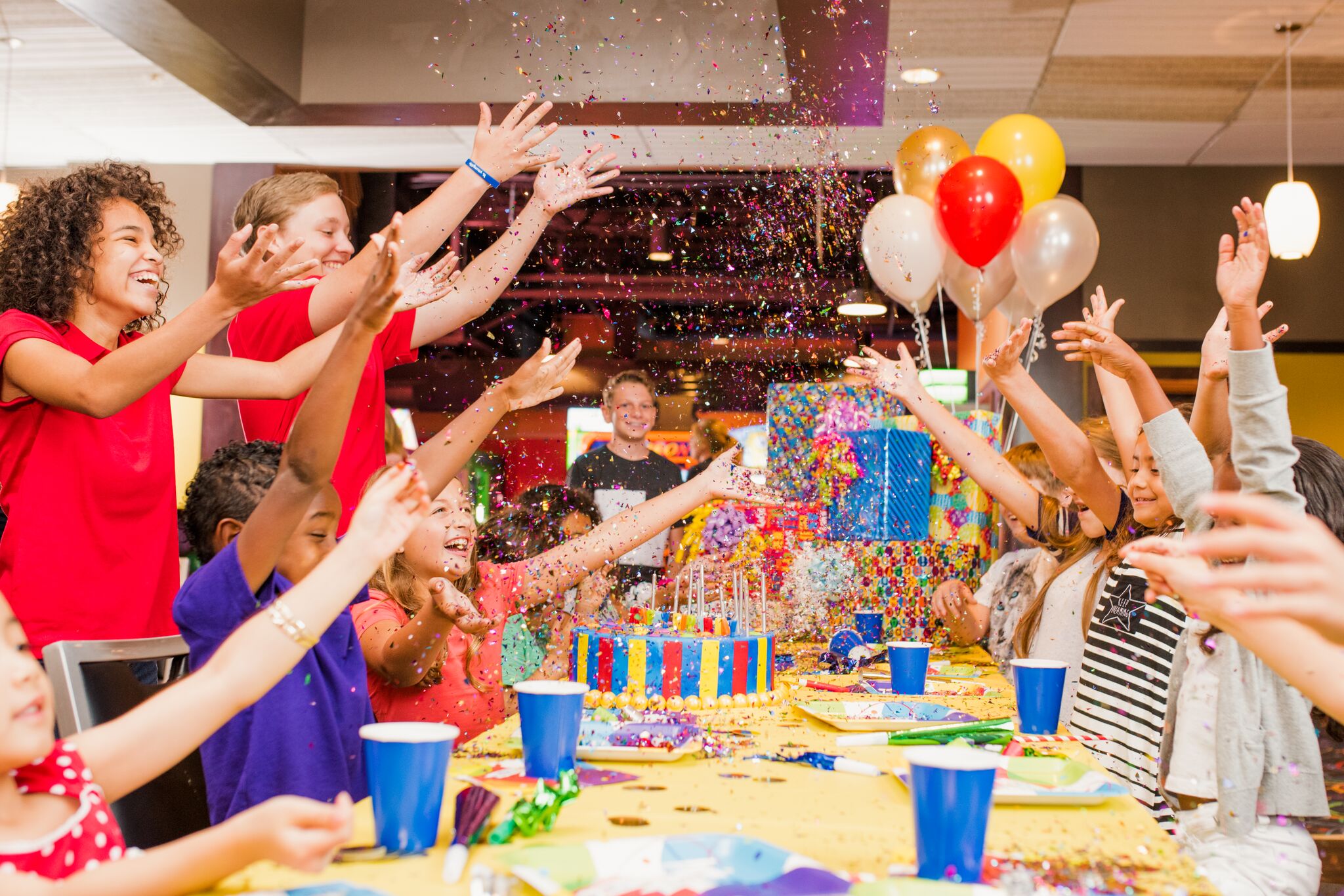 A group of happy children throwing colorful confetti over a birthday cake at a party table.