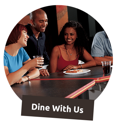 A group of four friends laughing and eating pizza together behind a Dine With Us banner.