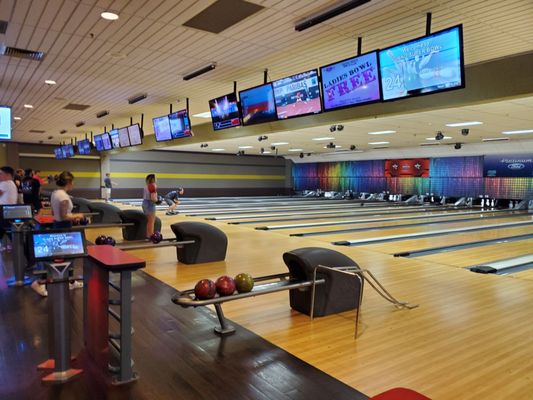 Wide view of a brightly lit bowling center with many lanes, overhead monitors, and bowlers on the approach.