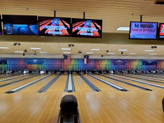 A wide view of numerous bowling lanes under bright lights with overhead scoring monitors and national and state flags.