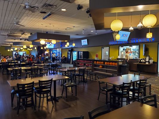 A large dining area with numerous tables and chairs next to a kitchen service counter and bowling ball storage racks.