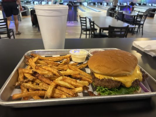 A cheeseburger with melted cheese and a pile of french fries on a metal tray inside a bowling alley.