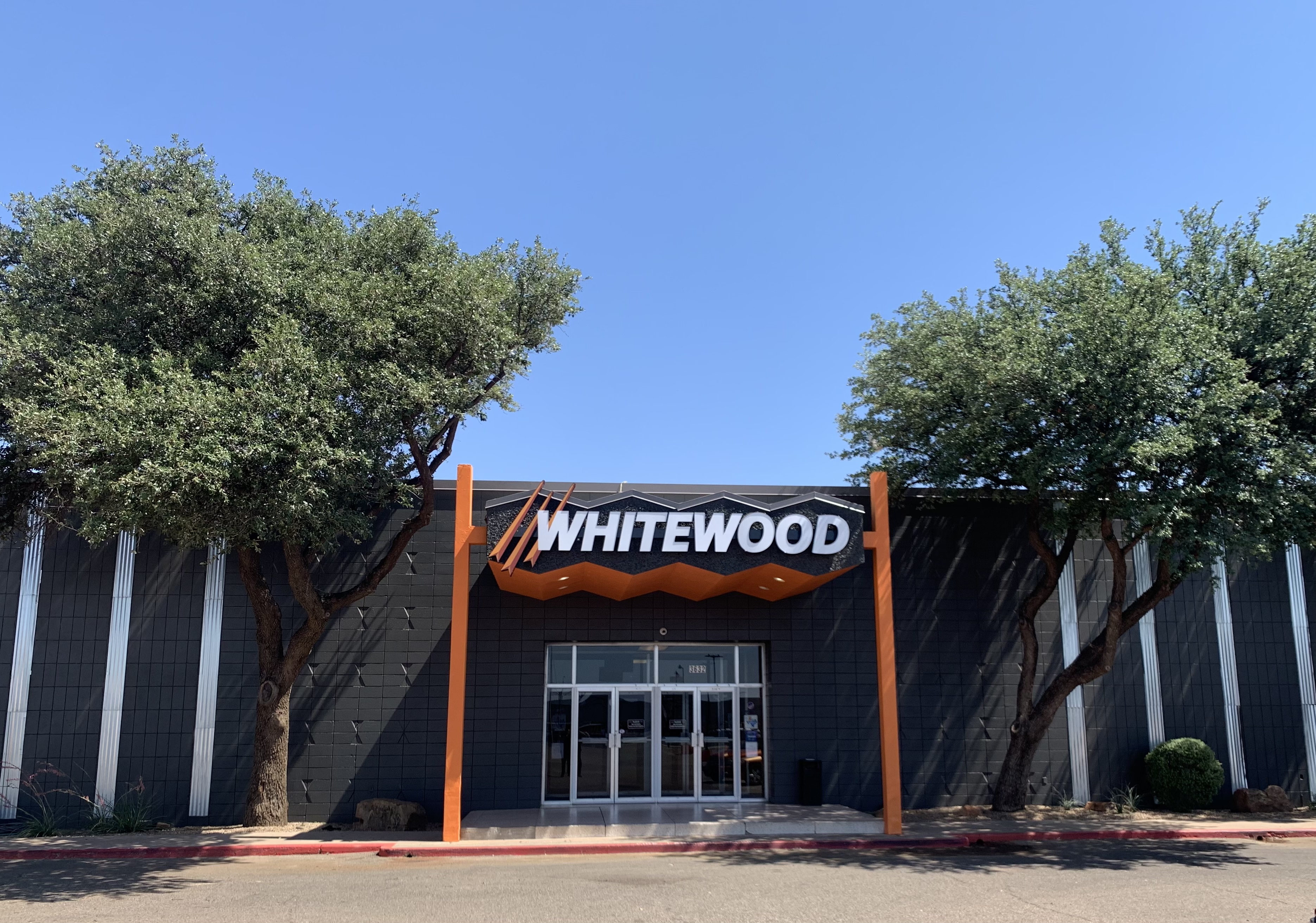 Exterior view of Whitewood Lanes building entrance with large "WHITEWOOD" logo under a clear blue sky, flanked by two trees.
