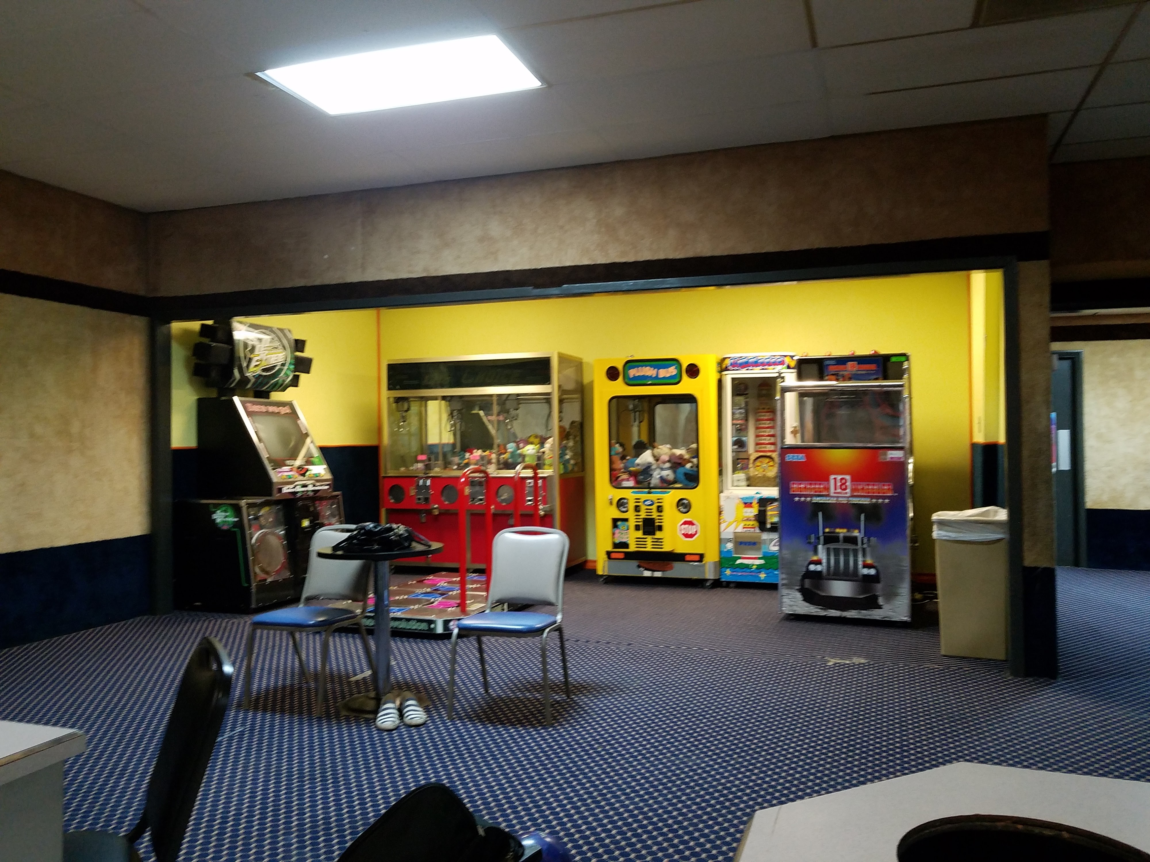 An arcade area with colorful claw machines, a rhythm game, and seating with tables on patterned blue carpet.