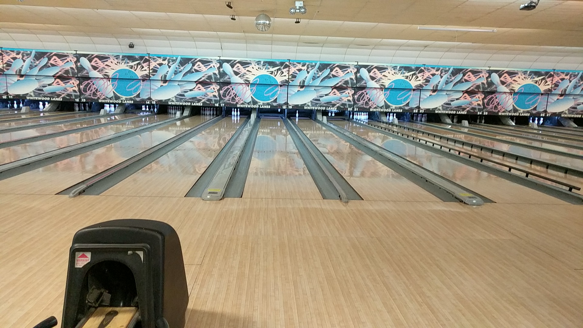 A wide shot of multiple empty bowling lanes at Whitewood Lanes with a colorful graphic mural above the pin decks.