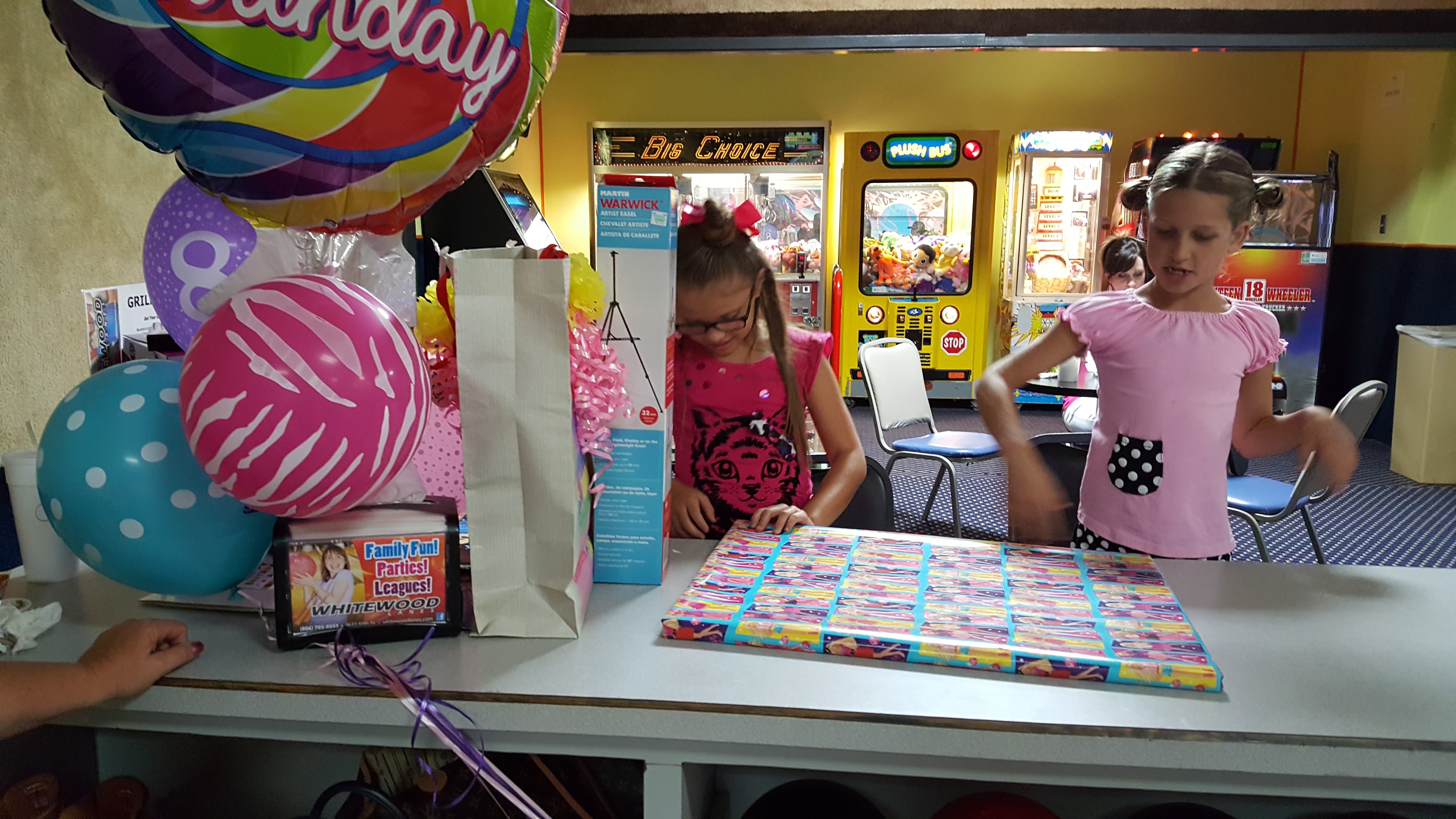 Two girls stand by a counter with birthday balloons and a gift, in front of several arcade machines.