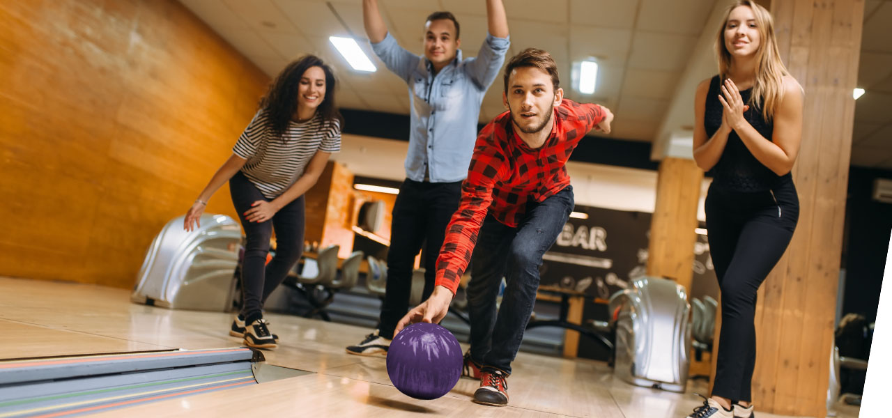 Four friends bowling, with one man actively throwing a purple bowling ball down the lane.