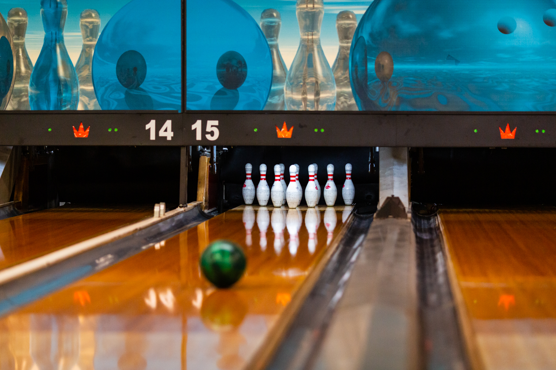 A bowling ball rolls down a polished wooden lane toward a set of ten white pins in a bowling center.