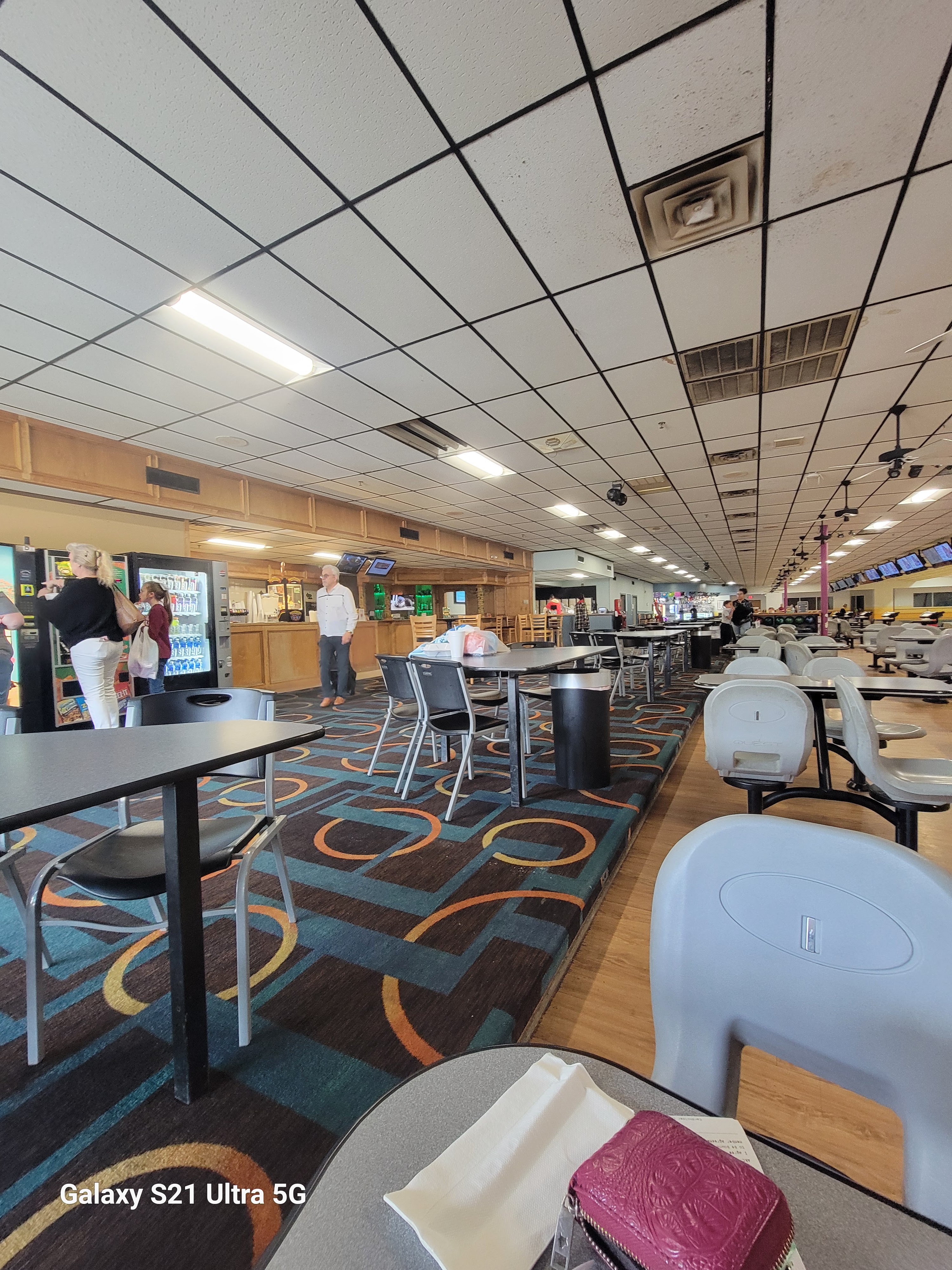 A bowling center concourse with tables, patterned carpet, vending machines, and a snack bar counter.