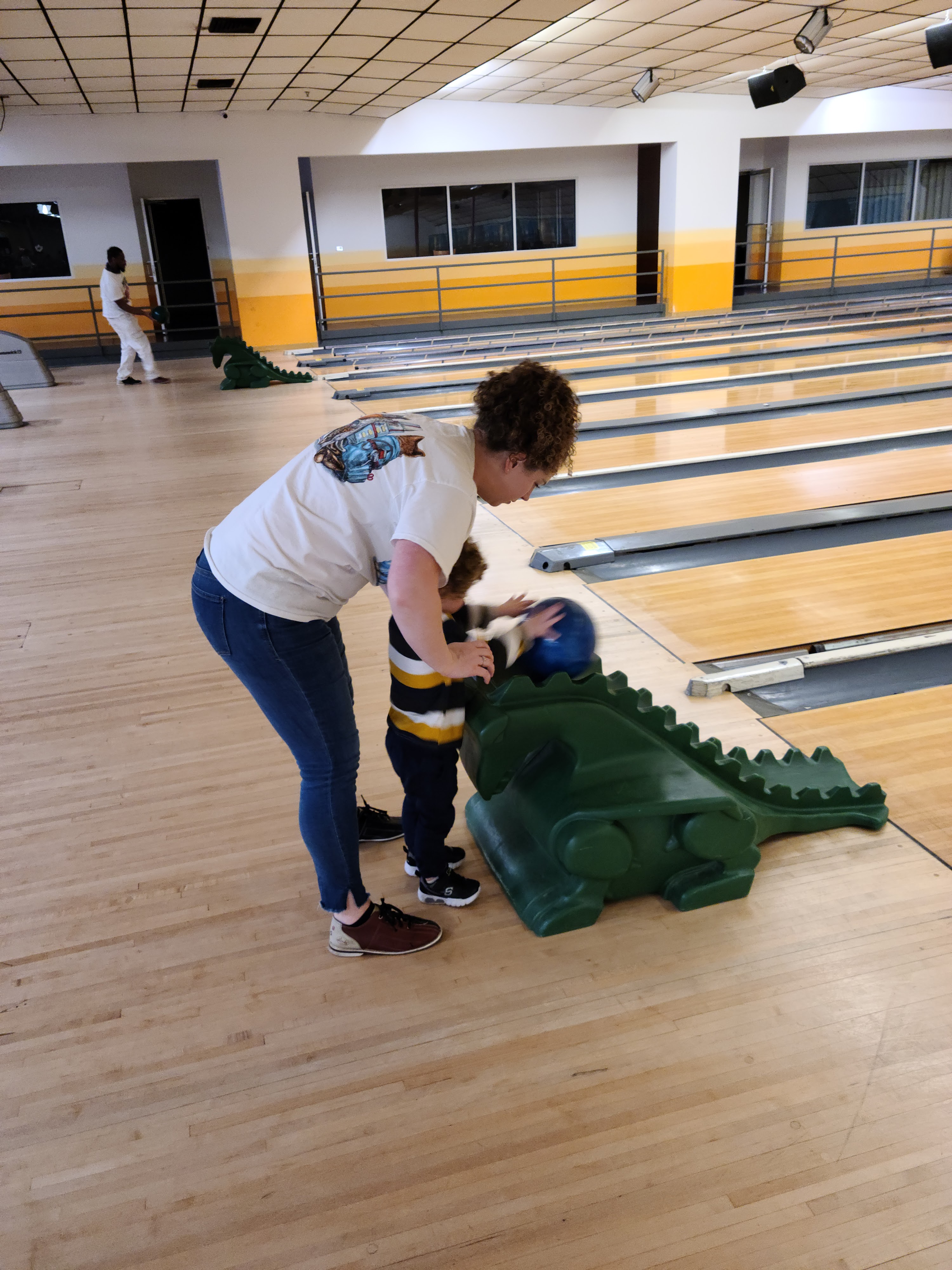 A woman helps a young child position a blue bowling ball on a green dinosaur-shaped ramp.
