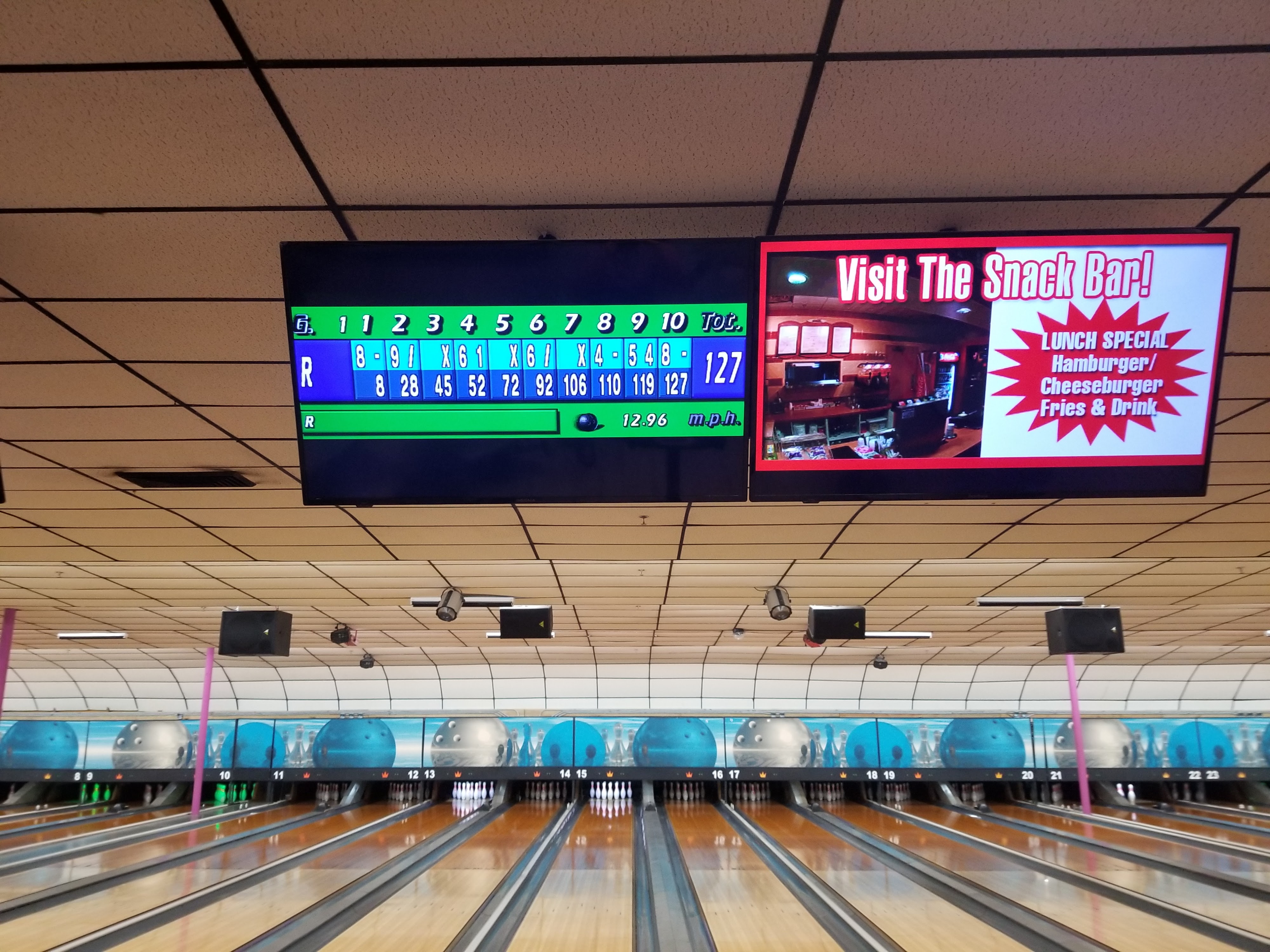 A view of several bowling lanes with overhead monitors showing a game score and a snack bar lunch special advertisement.