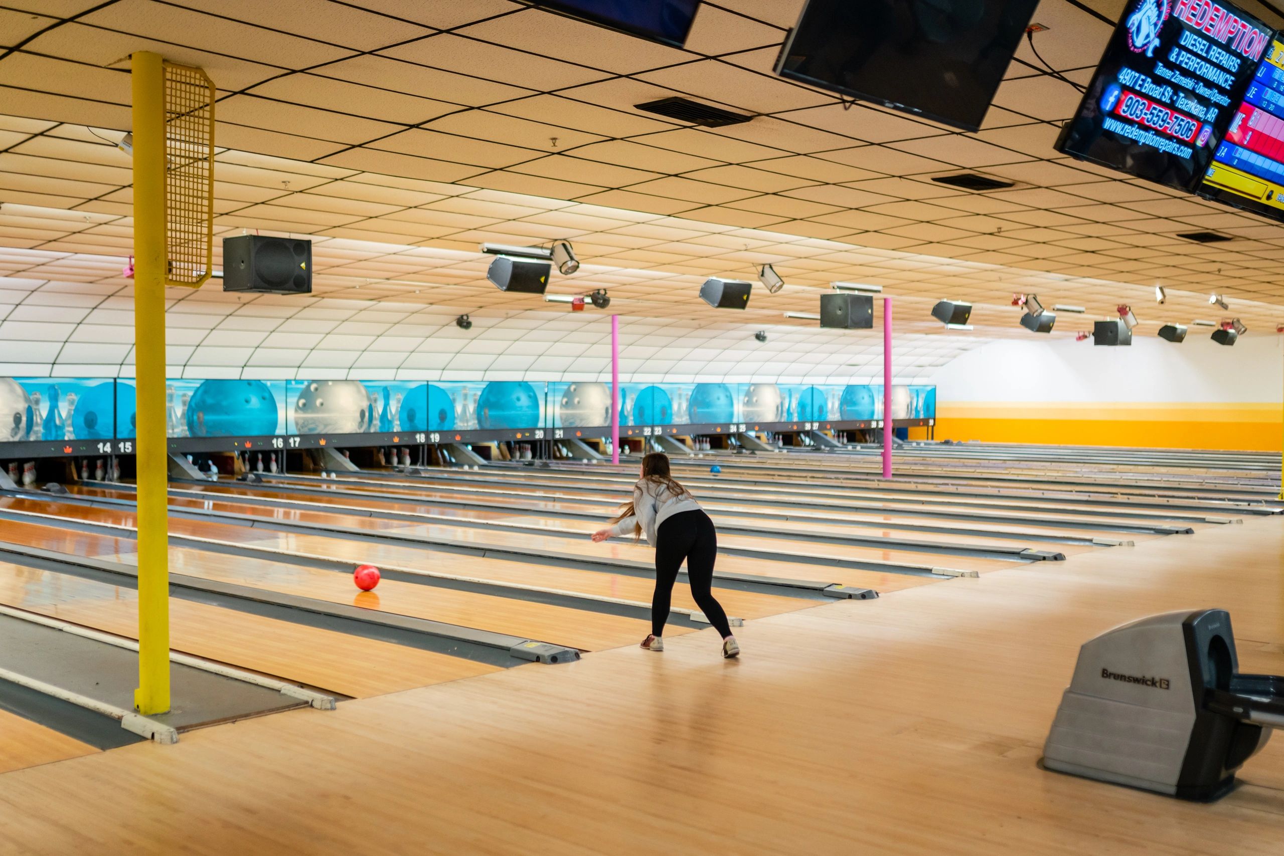 A woman in a grey sweatshirt bowls a red ball down a wooden lane in a bright bowling center.