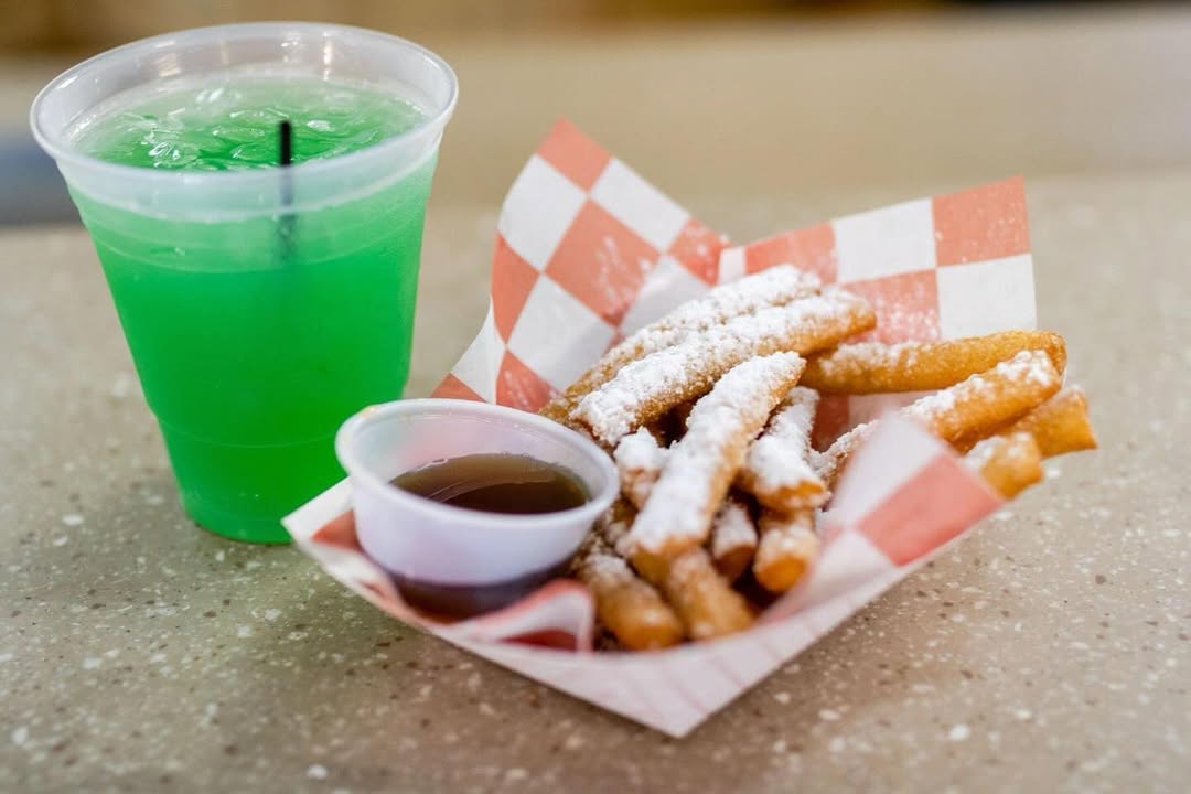 A tray of powdered sugar funnel cake fries with dipping sauce and a bright green iced drink on a table.