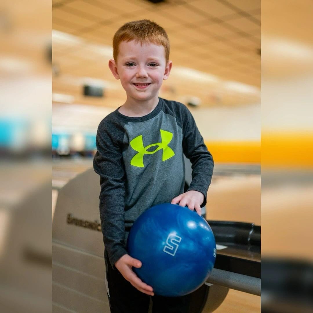 A young boy with red hair smiling and holding a blue bowling ball next to a bowling ball return.