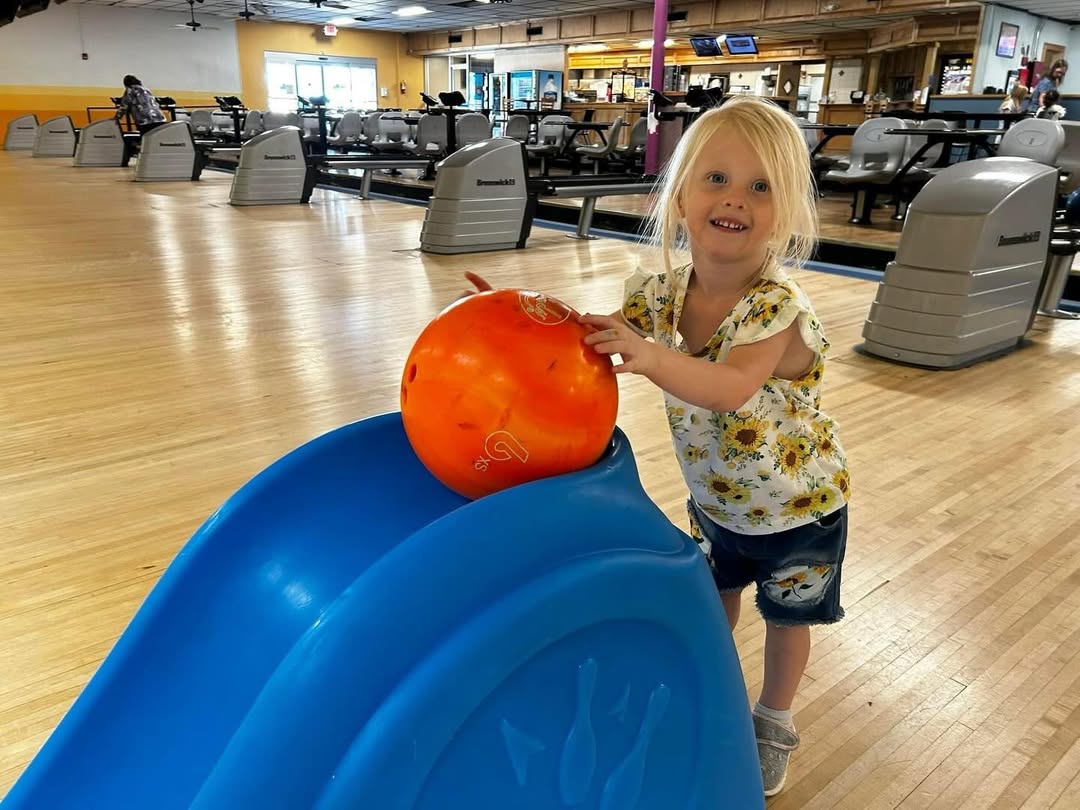 A smiling young girl uses a blue bowling ramp to roll an orange ball down the lane.