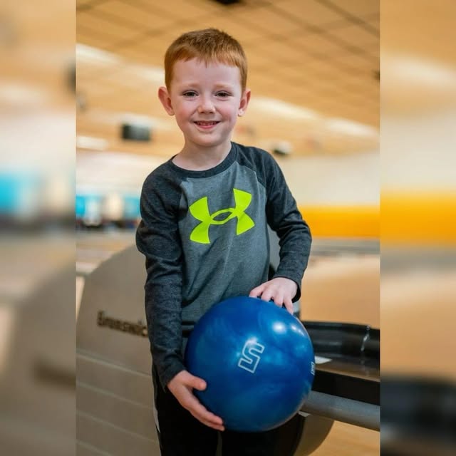 A young boy with red hair smiles while holding a blue bowling ball in a bowling alley.