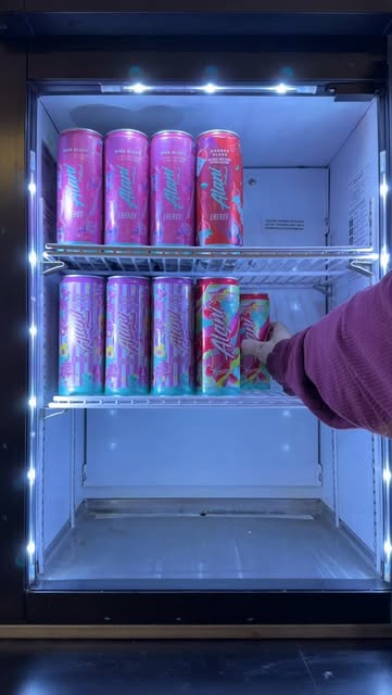 A hand reaches into a refrigerator filled with pink and colorful Alani Nu energy drink cans.