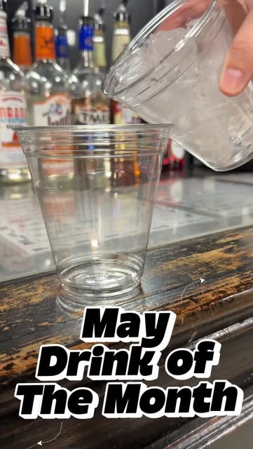 A hand pours ice from one clear cup into another clear cup on a wooden bar top. Liquor bottles are blurred in the background.