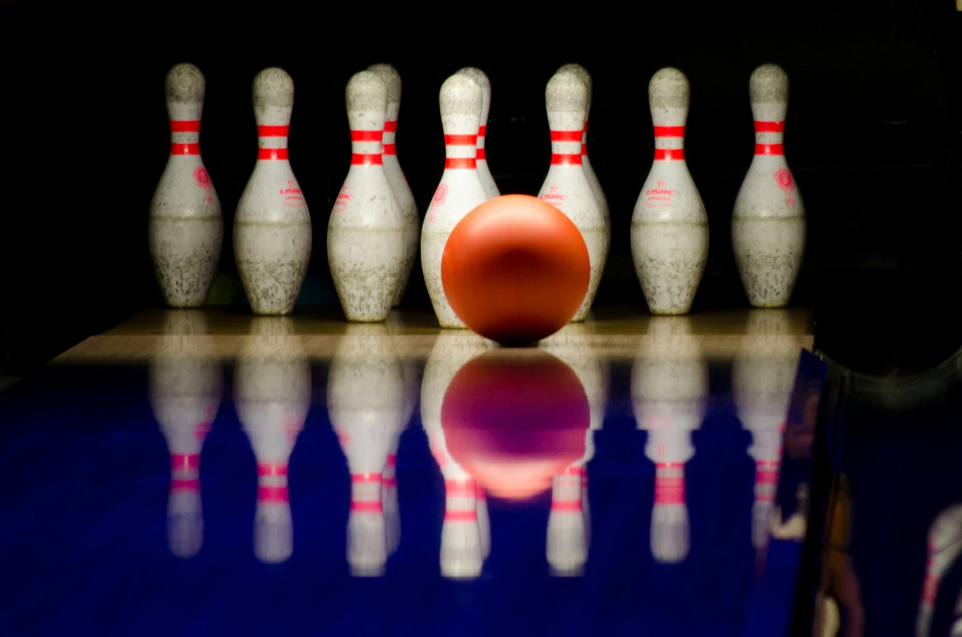 A red bowling ball rolls down a dark lane with blue reflections toward ten white bowling pins.