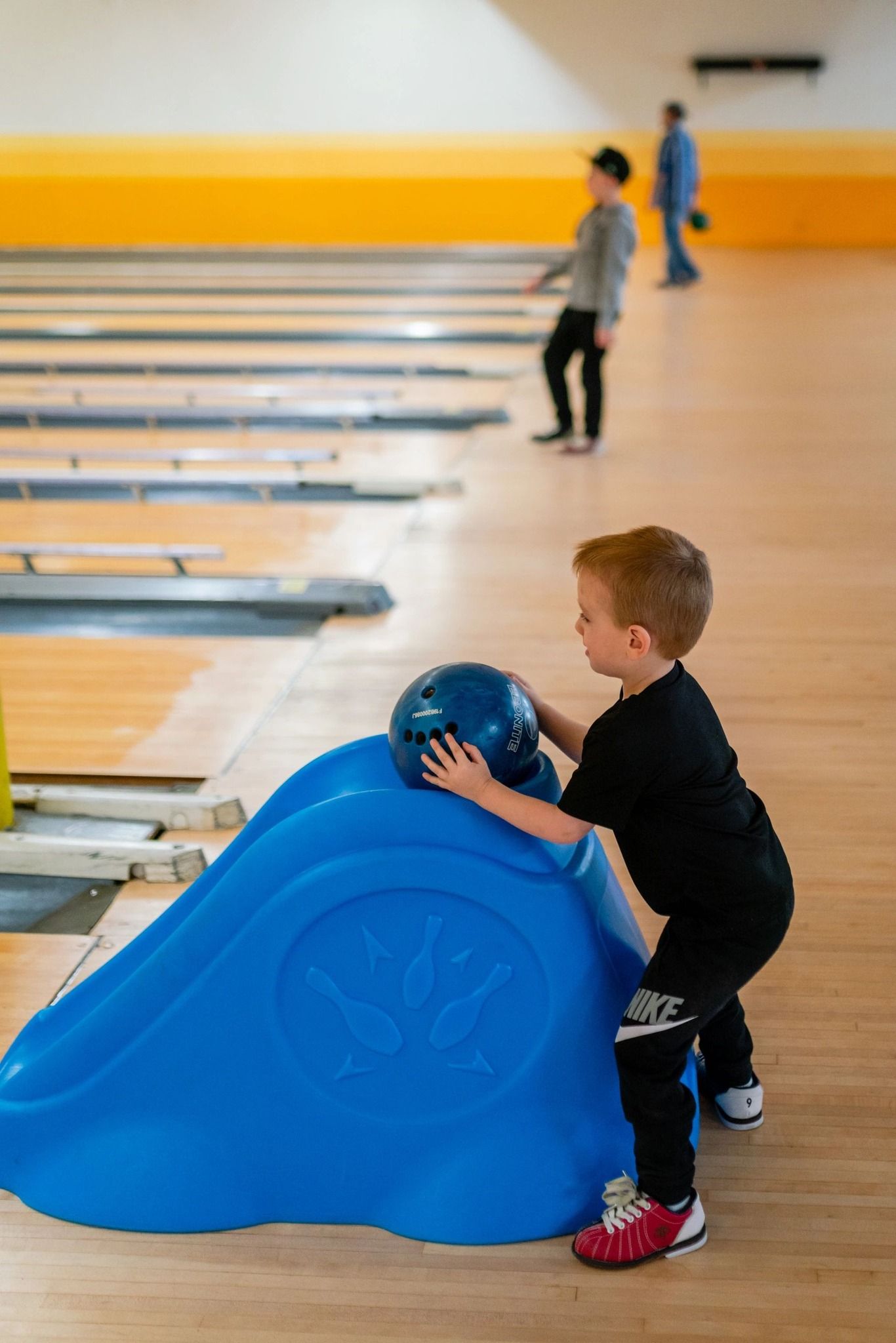 A young child using a blue bowling ramp to aim a ball down a wooden lane.