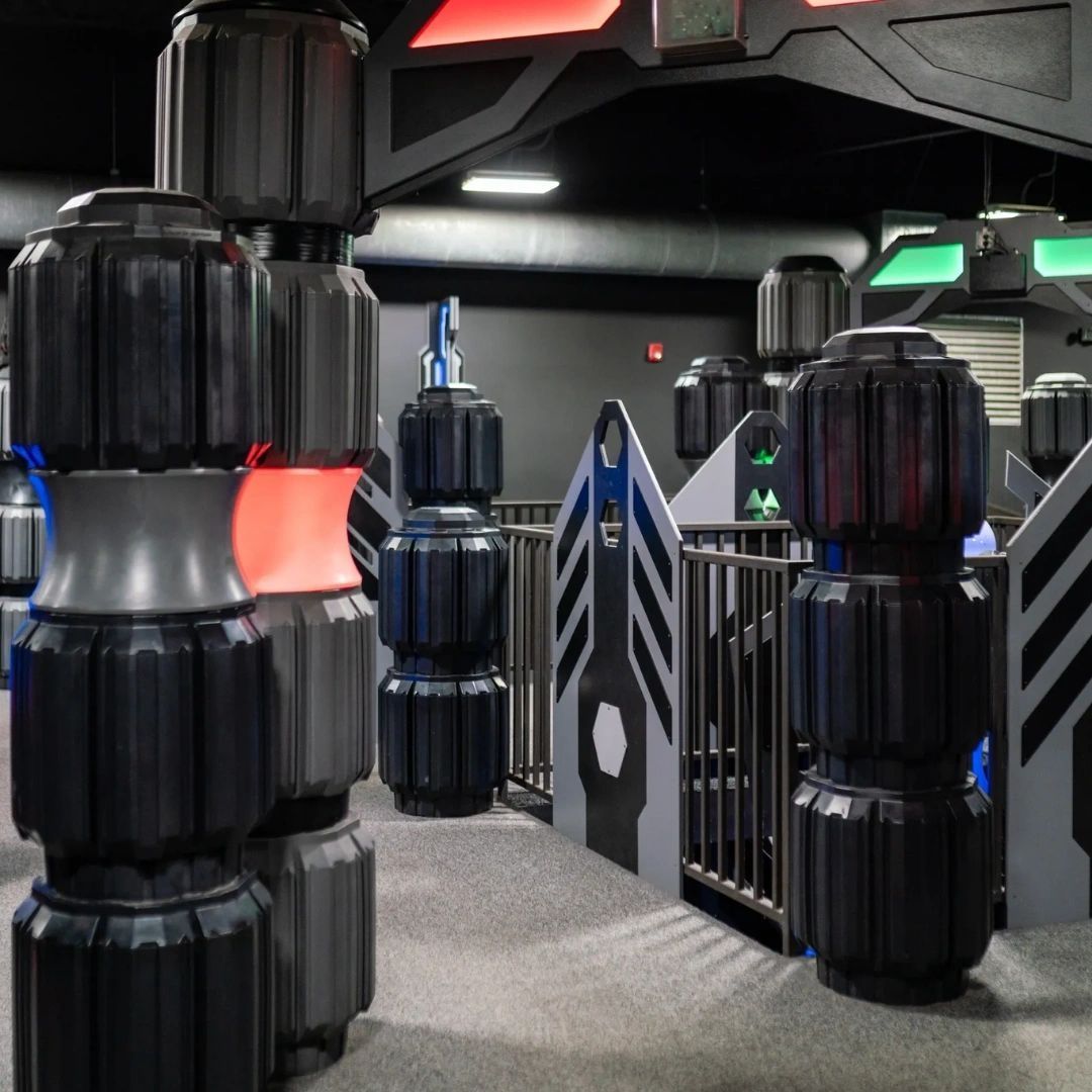 A modern indoor laser tag arena with glowing red and blue textured pillars and tactical wall barriers.
