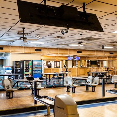 Interior view of a bowling alley showing seating, ball returns, and a snack bar area under bright ceiling lights.