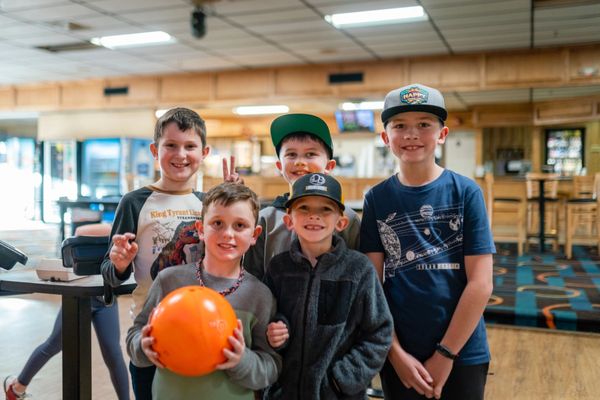 Five young boys smiling and posing together in a bowling alley, with one boy holding an orange bowling ball.